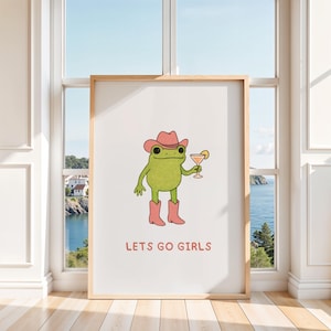 Könnte beinhalten: Ein gerahmter Kunstdruck mit einer Cartoon-Froschfigur, die einen rosa Cowboyhut und Stiefel trägt und einen Cocktail hält. Der Text "LETS GO GIRLS" ist darunter in Rosa gedruckt. Das Kunstwerk wird in einem Raum mit einem Fenster mit Blick auf eine Küstenlandschaft ausgestellt.