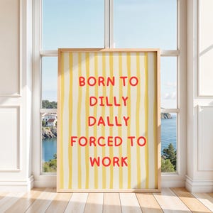 以下が含まれることがあります： 黄色と白のストライプの背景に、赤い文字で「BORN TO DILLY DALLY FORCED TO WORK」と書かれた額入りのアートプリント。 海岸の景色を望む窓のある部屋に飾られています。