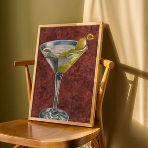May include: A framed rectangular art print featuring a watercolour illustration of a martini glass filled with a clear liquid and garnished with two green olives on a cocktail stick, set against a textured, deep maroon background.