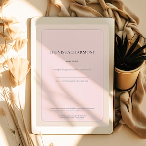 May include: A digital tablet displays a mini guide titled "THE VISUAL HARMONY" with text about design lessons for creative souls. The screen has a pink background with text in black. The tablet is on a beige surface with a plant in a gold pot.