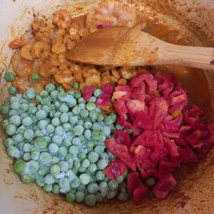Könnte beinhalten: Ein heller Kochtopf mit einer Mischung aus kleinen orangefarbenen Garnelen, gefrorenen grünen Erbsen und gewürfelten roten Paprika. Ein Holzlöffel liegt auf den Zutaten, die in dem Topf mit einer rotbraunen Soße bedeckt sind.