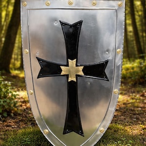 Puede incluir: Un escudo plateado con una cruz negra y una cruz dorada en el centro. El escudo tiene una parte superior redondeada y está bordeado con remaches dorados. El fondo es una escena de bosque borrosa.