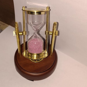Puede incluir: Un reloj de arena con una cámara de vidrio transparente llena de arena rosa, sostenido por un marco de metal dorado. El reloj de arena se asienta sobre una base redonda de madera oscura. El diseño incluye detalles metálicos dorados.