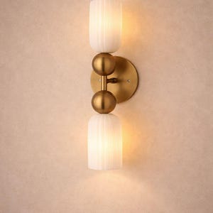 Può includere: Applique da parete moderno con finitura in ottone e due paralumi in vetro bianco a coste. L'applique ha un design verticale con una piastra rotonda e due accenti sferici. Apparecchio di illuminazione per interni.