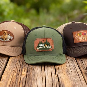 Könnte beinhalten: Drei Trucker-Caps mit Mesh-Rücken auf einer Holzoberfläche. Eine ist beige mit einem Hunde- und Katzenaufnäher. Die zweite ist grün mit einem "Summer Camp"-Aufnäher. Die dritte ist braun mit einem "Logo in Here"-Aufnäher.
