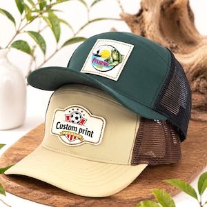 Op de afbeelding: Twee baseballpetten op elkaar gestapeld. De bovenste pet is donkergroen met een zwart mesh-achterpand en een patch met de tekst "Fishing Club" met een visafbeelding. De onderste pet is beige met een bruin mesh-achterpand en een patch met de tekst "Custom print".