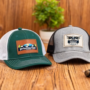 Peut inclure: Deux casquettes de camionneur avec des dos en filet sont présentées. L'une est vert foncé avec un dos en filet blanc et un patch en cuir avec le texte "printing logo". L'autre est grise avec un dos en filet noir et un patch "Eagles".