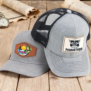 Puede incluir: Dos gorras de béisbol grises con diferentes parches. Una tiene un parche con temática de pesca con un pescador, un pez y una puesta de sol. La otra tiene una parte trasera de malla negra y un parche con un logotipo de águila y las palabras "Printed logo".