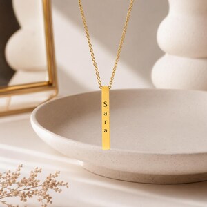 Op de afbeelding: Een goudkleurige verticale staafhanger aan een ketting met de naam Sara gegraveerd in zwarte letters. De hanger hangt aan een fijne goudkleurige ketting en heeft een strak, langwerpig rechthoekig ontwerp met een gepolijste metalen afwerking.