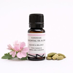 Puede incluir: Una botella de vidrio marrón de aceite esencial con tapa negra, etiquetada como "Handmade Essential Oil Blend Bloom & Balance". La imagen incluye flores rosas, hojas verdes y vainas de cardamomo. La botella mide aproximadamente 7.6 cm de alto.