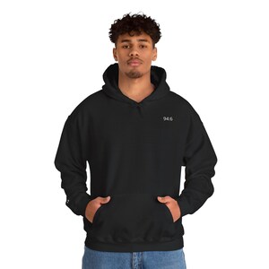 May include: A black hooded sweatshirt with a front pocket and the text "94:6" in white on the upper chest. The sweatshirt is worn by a person. The person is wearing blue jeans.