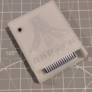 May include: A white 3D-printed cartridge case for an Atari game. The case features the Atari logo and the text "Atari 2600". The cartridge slot reveals a blue and silver connector.