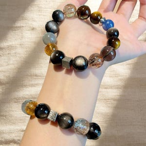 May include: Two beaded bracelets featuring a variety of colorful, spherical gemstones. The bracelets include black, brown, yellow, and iridescent beads, along with a silver-colored square accent. The bracelets are displayed on a person's hand.