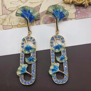 Pode incluir: Um par de brincos pendentes dourados com um topo em forma de folha de ginkgo e uma moldura oval alongada. A moldura é contornada por pequenos cristais azuis e contém três pingentes de folha de ginkgo com acabamento em esmalte azul e verde.