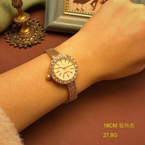 May include: A gold-tone wristwatch with a round face, a bezel set with clear crystals, and a fine mesh metal band. The watch face is cream-colored with gold-tone hands and markers. The text 18CM and 27.8G is visible in the lower corner.