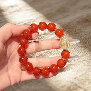 May include: A beaded bracelet featuring large, round, reddish-orange beads. The bracelet includes a single, larger, translucent yellow bead and gold-colored spacers. The bracelet is held in a hand.