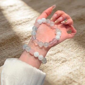 Könnte beinhalten: Zwei Perlenarmbänder bestehend aus einer Mischung aus durchscheinenden hellblauen, klaren und blassrosa kugelförmigen Perlen. Die Armbänder sind auf einem elastischen Band aufgezogen und weisen eine glatte, polierte Oberfläche mit unterschiedlicher Transparenz auf.