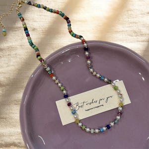 Puede incluir: Un collar de cuentas que alterna pequeñas perlas blancas y cuentas cuadradas multicolores en tonos azul, verde, rojo, amarillo y morado. Debajo hay una tarjeta rectangular con el texto impreso Best wishes for you, happiness is happening.