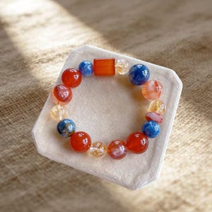May include: A beaded bracelet featuring a mix of round beads in shades of red, blue, and translucent yellow, along with a single rectangular orange bead. The beads are arranged in a circular pattern, showcasing varied textures and color gradients.