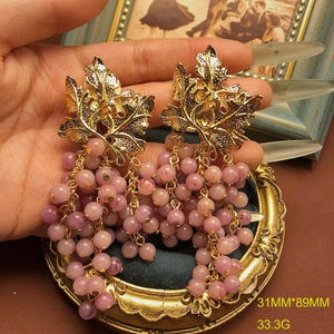 May include: A pair of gold-tone metal earrings featuring a textured grape leaf design at the top, with clusters of small, round, light purple beads hanging below. The earrings measure 31mm by 89mm and have a total weight of 33.3 grams.