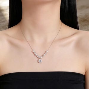 Pode incluir: Um colar delicado de tom prateado com um pingente central de pedra transparente de corte quadrado rodeado por um halo de pequenos cristais. A corrente é acentuada com pérolas brancas redondas e pequenos cristais em forma de V.