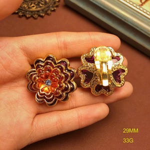 May include: A pair of gold-tone flower-shaped clip-on earrings featuring layered petals in shades of deep purple and orange with a small blue center stone. The earrings measure 29mm in diameter and have a total weight of 33g as indicated in text.