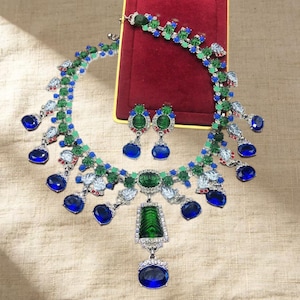 May include: A jewelry set consisting of a necklace and matching earrings. The necklace features green and blue faceted stones, clear crystals, and a central pendant with a large blue oval stone. The earrings mirror the necklace design.