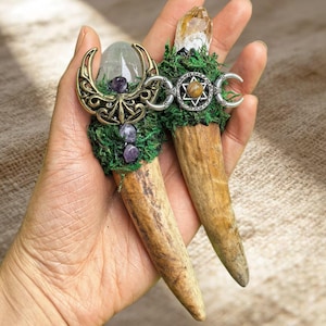 May include: Two decorative wands featuring natural antler bases topped with green moss. One wand has a clear crystal, purple stones, and a gold-toned crescent moon frame. The other has a citrine point, a silver-toned hexagram, and crescent moons.