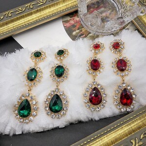 May include: Two pairs of drop earrings featuring faceted stones surrounded by clear crystals. One pair has deep green oval and teardrop-shaped stones, while the other pair features vibrant red stones in matching oval and teardrop shapes.