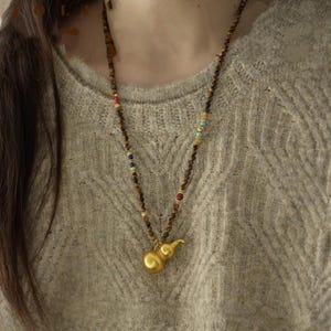 Peut inclure: Un long collier de perles avec un pendentif en forme de gourde de couleur or. Le collier est composé de petites perles rondes marron, entrecoupées de perles de couleur. Le pendentif en forme de gourde est lisse et brillant, avec une tige détaillée.