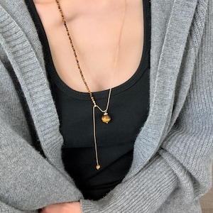 Puede incluir: Un collar dorado con dos cadenas. Una cadena presenta pequeñas cuentas marrones, mientras que la otra tiene un solo colgante redondo de ojo de tigre. Un pequeño dije de corazón dorado cuelga del extremo de la cadena más larga. El collar se usa sobre una camiseta negra.