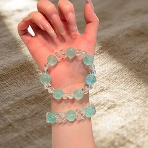 May include: Two bracelets, one on the wrist and one above, featuring clear and light blue beads. The light blue beads have a carved floral design. The bracelets are strung together with clear beads.