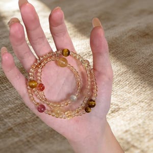 May include: Three beaded bracelets in shades of amber, gold, and clear. The bracelets feature a variety of gemstone beads, including tiger's eye, citrine, and other colorful stones. The bracelets are displayed on a hand.
