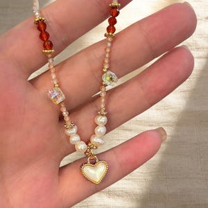 Puede incluir: Un collar de cuentas con un colgante central en forma de corazón blanco con borde dorado. La cadena incluye perlas de agua dulce blancas, cristales facetados translúcidos y cuentas de color naranja oscuro, acentuadas con pequeños espaciadores metálicos dorados.