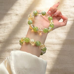 Peut inclure: Deux bracelets de perles. Un bracelet est au poignet, l'autre est soulevé. Les bracelets présentent des perles vertes et jaunes. Les perles sont rondes et facettées. Le fond est de couleur beige clair.