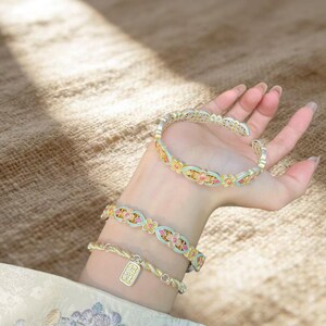 Pode incluir: Duas pulseiras abertas e uma pulseira de corrente com um motivo floral repetitivo em tons de azul claro, rosa e dourado. A pulseira de corrente inclui um pequeno pingente retangular com detalhes gravados, tudo trabalhado em metal.