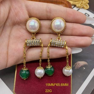 Peut inclure: Une paire de boucles d'oreilles pendantes dorées avec un clou en perle irisée, un cylindre horizontal incrusté de cristaux transparents et deux chaînes pendantes terminées par une perle verte et une perle blanche, mesurant 15mm par 65,6mm et 22g.