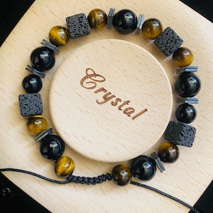 Puede incluir: Una pulsera de cuentas con ónix negro, ojo de tigre y piedra de lava. Incluye pequeños espaciadores grises rectangulares y está ensartada en un cordón negro. La palabra "Crystal" está grabada en la superficie de madera.