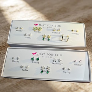 May include: Two white rectangular boxes labeled JUST FOR YOU BEST WISHES containing seven pairs of earrings each, organized by days of the week. The earrings feature various designs including small studs, pearls, green stones, and clear crystals.