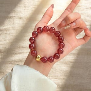 May include: A beaded bracelet featuring translucent red beads and a gold-colored accent bead. The bracelet is displayed on a person's wrist, showcasing its color and design. The beads appear to be approximately 0.5 inches in diameter.