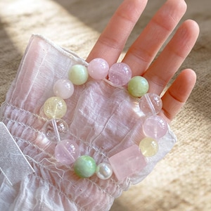 May include: A stretch bracelet featuring a variety of smooth, round beads in shades of pale pink, light green, yellow, and clear, along with one rectangular pink bead and a single small white pearl, arranged in a circular pattern on a wrist.