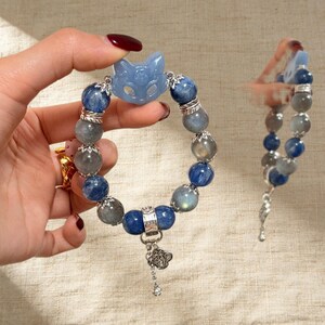 May include: A beaded bracelet featuring blue and gray gemstone beads, a light blue cat-shaped charm, and silver accents. The bracelet is held up, showcasing its design and details. A second bracelet is also visible.