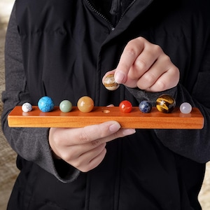 May include: A rectangular wooden display stand featuring a row of circular indentations holding various polished gemstone spheres in colors including white, blue, green, orange, red, dark blue, brown, and translucent pink, with one sphere held above.