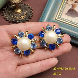 May include: A pair of gold-tone earrings featuring a central cream-colored faux pearl surrounded by alternating light blue and dark blue round crystals in a sunburst arrangement. The earrings measure 29.4mm by 29.4mm and have a weight of 26.7g.