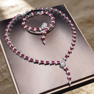 May include: A jewelry set consisting of a necklace and a bracelet, both featuring a snake-inspired design. The pieces are crafted with silver-toned metal, set with rectangular red stones and small clear white stones along the entire length.