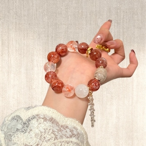 Könnte beinhalten: Ein Armband mit großen, runden Perlen in Rot-, Weiß- und transparenten Farbtönen. Es hat einen goldfarbenen Verschluss und einen Anhänger mit klaren, facettierten Perlen. Das Armband wird an einem Handgelenk gezeigt. Die Perlen haben einen Durchmesser von ca. 1,5 cm.
