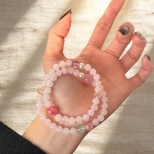 Könnte beinhalten: Zwei zarte Rosenquarz-Perlenarmbänder mit Akzentperlen in Rosa-, Lila- und Gelbtönen. Die Armbänder werden an einem Handgelenk getragen und zeigen den zarten Rosaton der Steine. Ein schönes Accessoire.