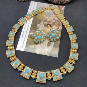 Könnte beinhalten: Eine goldfarbene Metallhalskette und ein passendes Paar runder Ohrstecker, jeweils mit eingebetteten runden türkisfarbenen Cabochons. Die Halskette besteht aus sich wiederholenden quadratischen Gliedern, die durch kleinere zylindrische Abstandshalter verbunden sind.