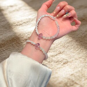 May include: Two bracelets featuring small white pearls and translucent, faceted light purple beads. Each bracelet includes a single, larger, irregular-shaped pink crystal bead as a central focal point, connected by a silver-toned metal clasp.