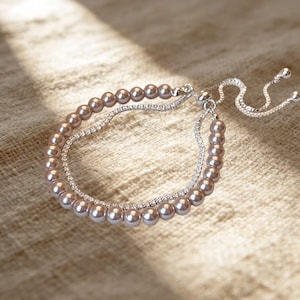 May include: A double-strand bracelet featuring one row of light brown spherical beads and one row of clear, sparkling stones set in silver-toned metal. The bracelet includes an adjustable sliding clasp mechanism with two dangling silver chains.